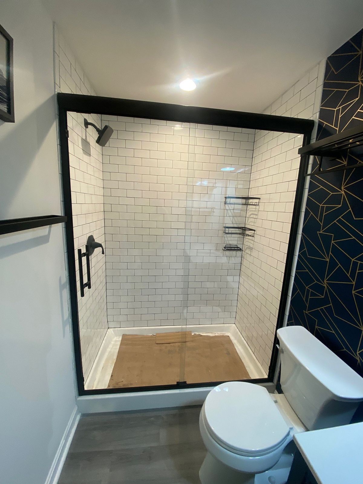 A walk-in shower with white subway tiles, a black frame, and a tan floor mat, next to a white toilet in a modern bathroom.