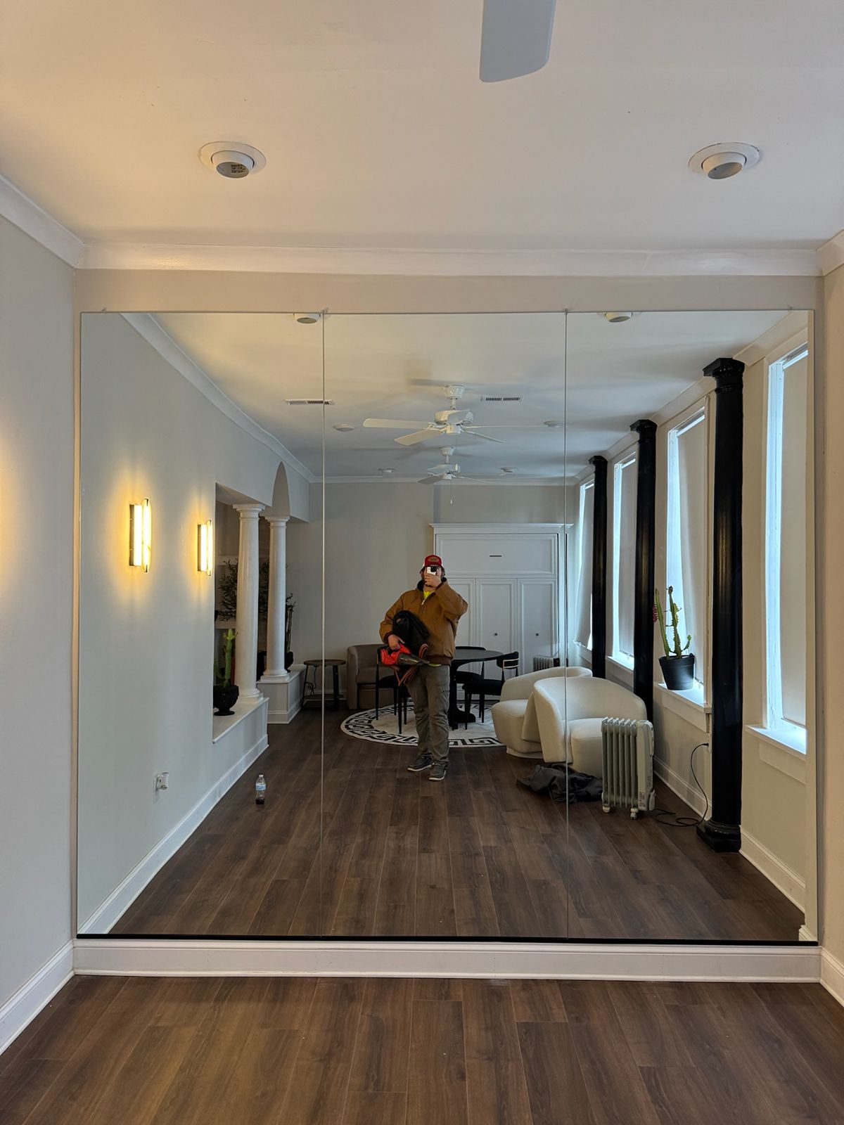A person in a brown jacket stands in front of a large wall mirror, reflecting a furnished living room with white chairs.