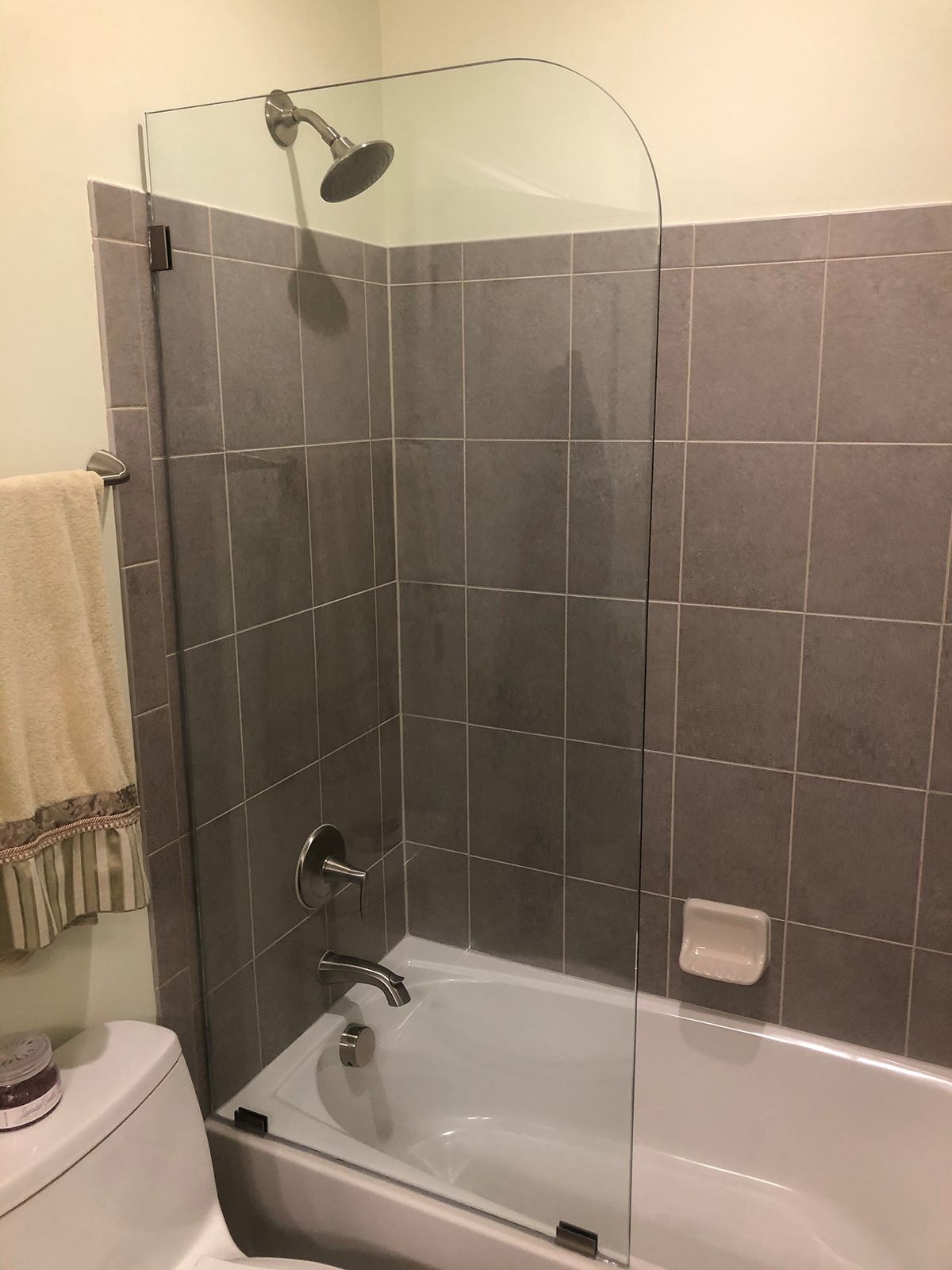 A bathtub with a gray tiled wall, a chrome shower fixture, and a glass partition door. A towel hangs nearby on the left.