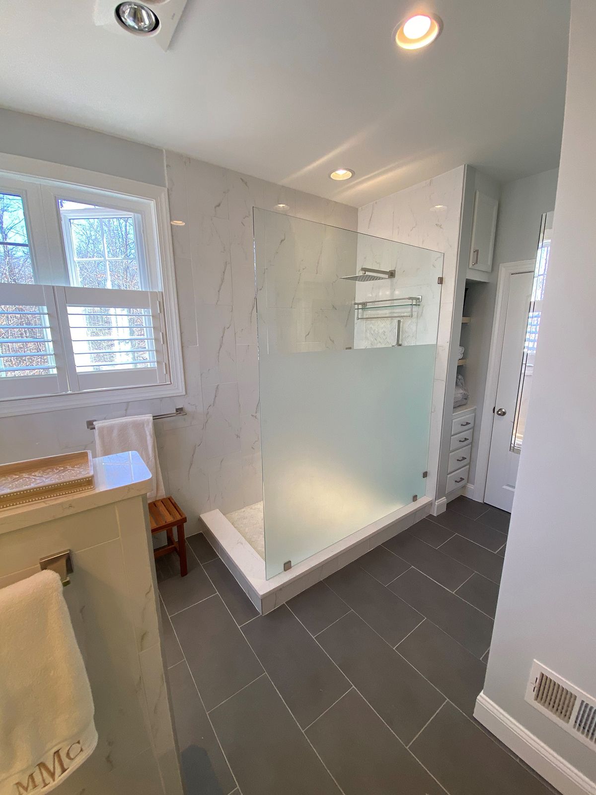A modern bathroom with grey floor tiles, a marble-tiled walk-in shower with frosted glass, and a window with white shutters.