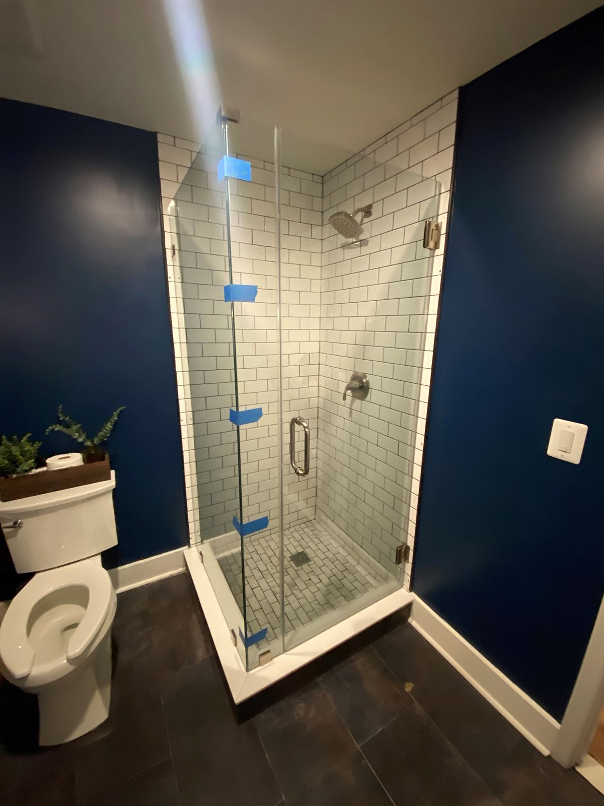 A modern bathroom with a glass-enclosed shower with white subway tiles, dark blue walls, and a white toilet on the left.