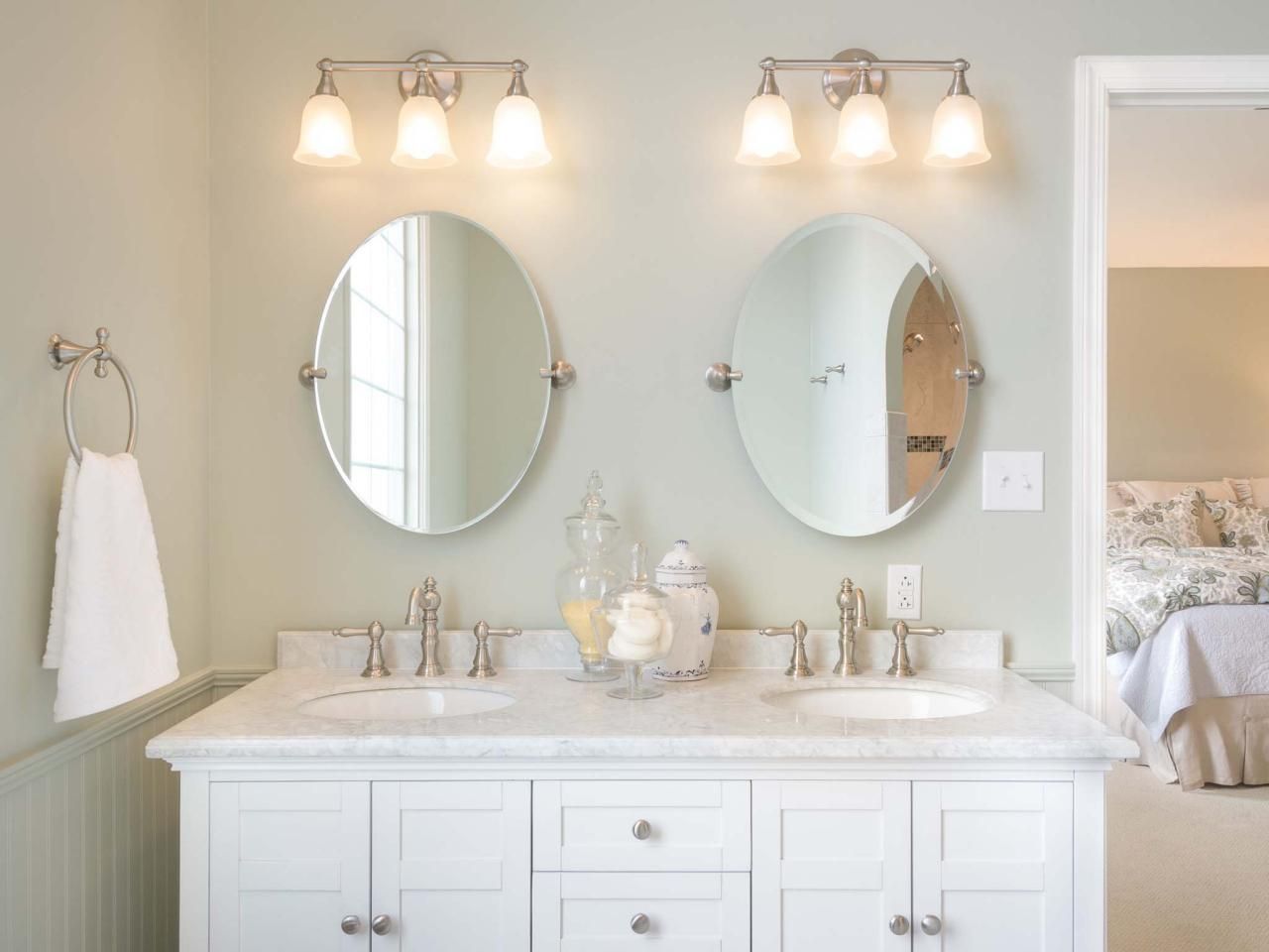 Bathroom with white vanity, oval mirrors, and light fixtures above sinks. A doorway leads to a bedroom.