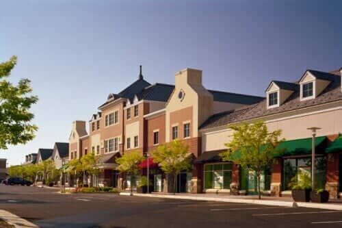 Shops and businesses line a street with a clear blue sky above. Trees and parked cars are visible.