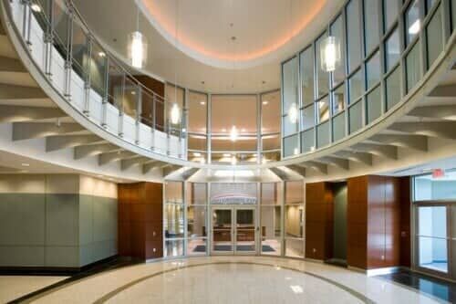 Modern building interior with curved glass walls, upper level balcony, and tile floor.