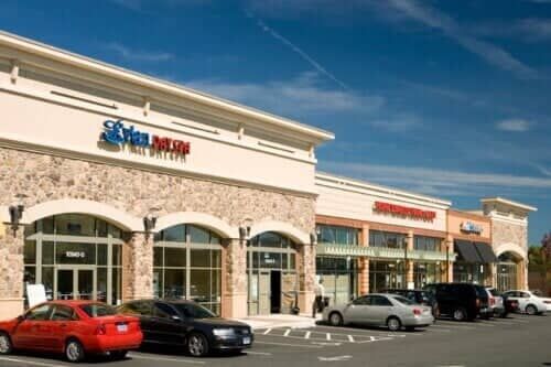 A row of shops in a strip mall with parked cars on a sunny day.