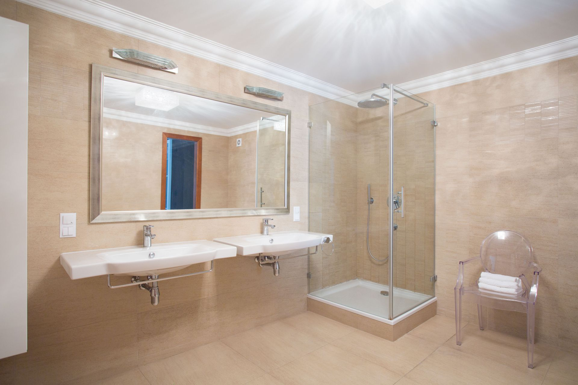 Bathroom with dual sinks, large mirror, glass shower, and clear chair. Beige tile and walls, bright lighting.