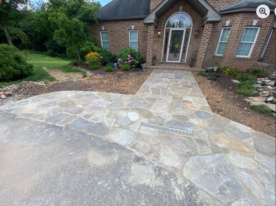 Stone pathway leading to a brick house with landscaping. Gray flagstones curve towards a front door, with a drainage grate visible.