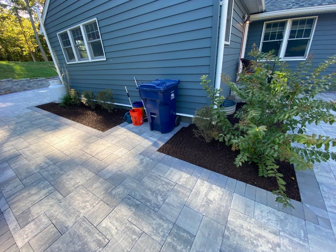 Paver patio alongside a blue house with landscaping beds, a blue trash can, and an orange bucket.