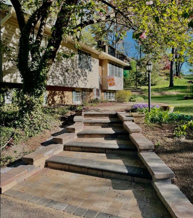 Stone steps lead up to a light-colored building with a tree canopy overhead. A lamppost and landscaping flank the steps.