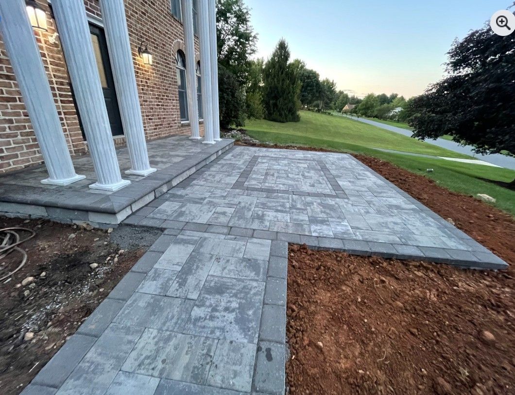 Stone patio and walkway leading to a brick house with white columns.  The area is surrounded by bare soil and a grassy hill.