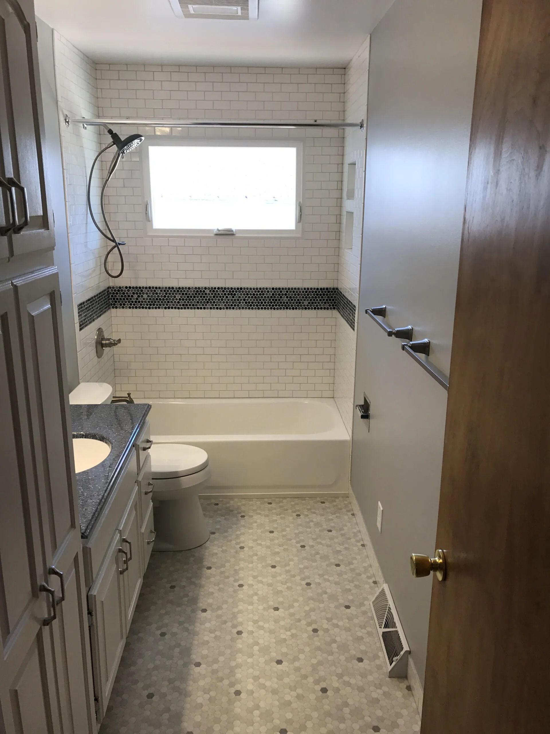 Narrow bathroom with white tile, bathtub, toilet, window, and wooden door partially open.