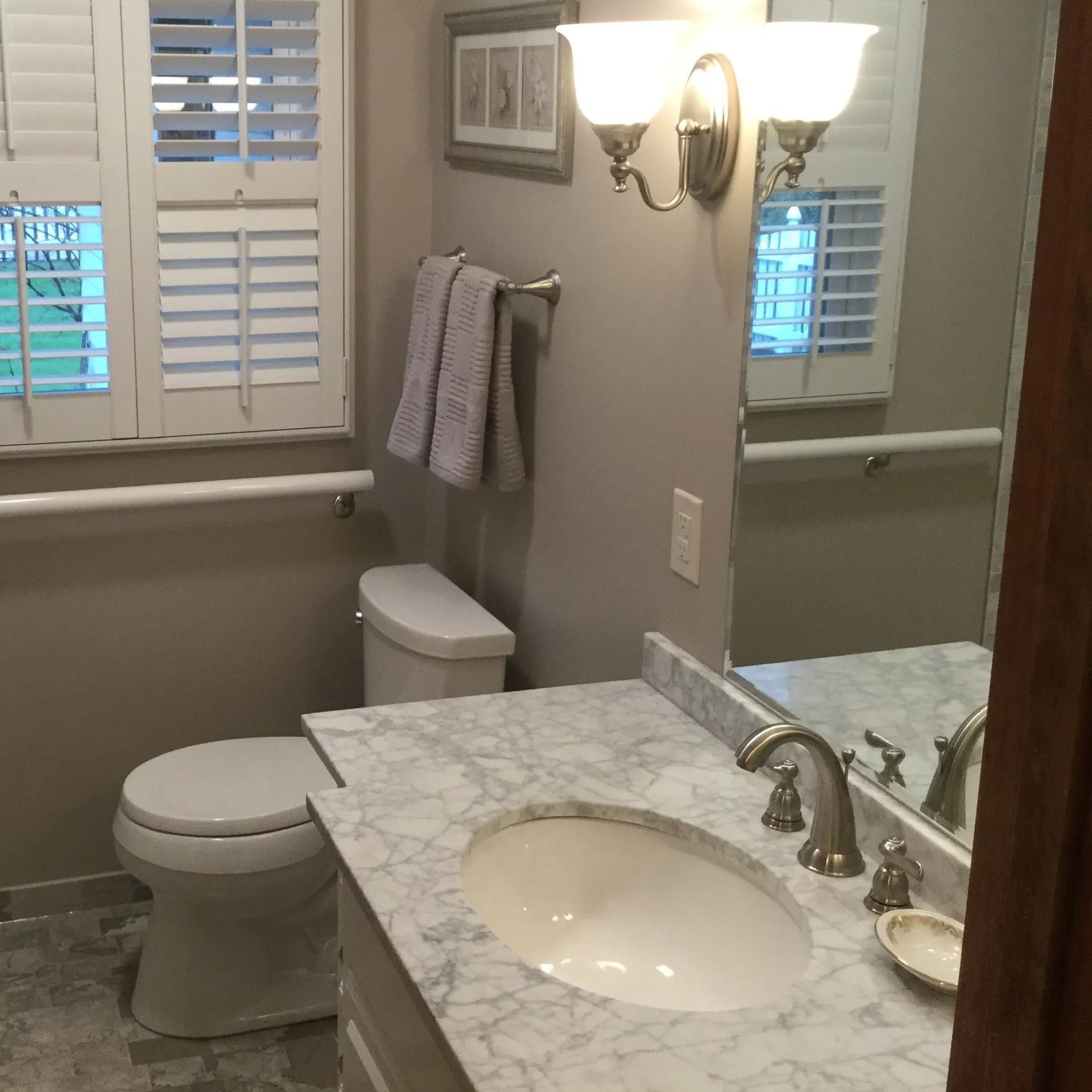 Bathroom with marble vanity, sink, toilet, mirror, and wall sconces beside a window with shutters