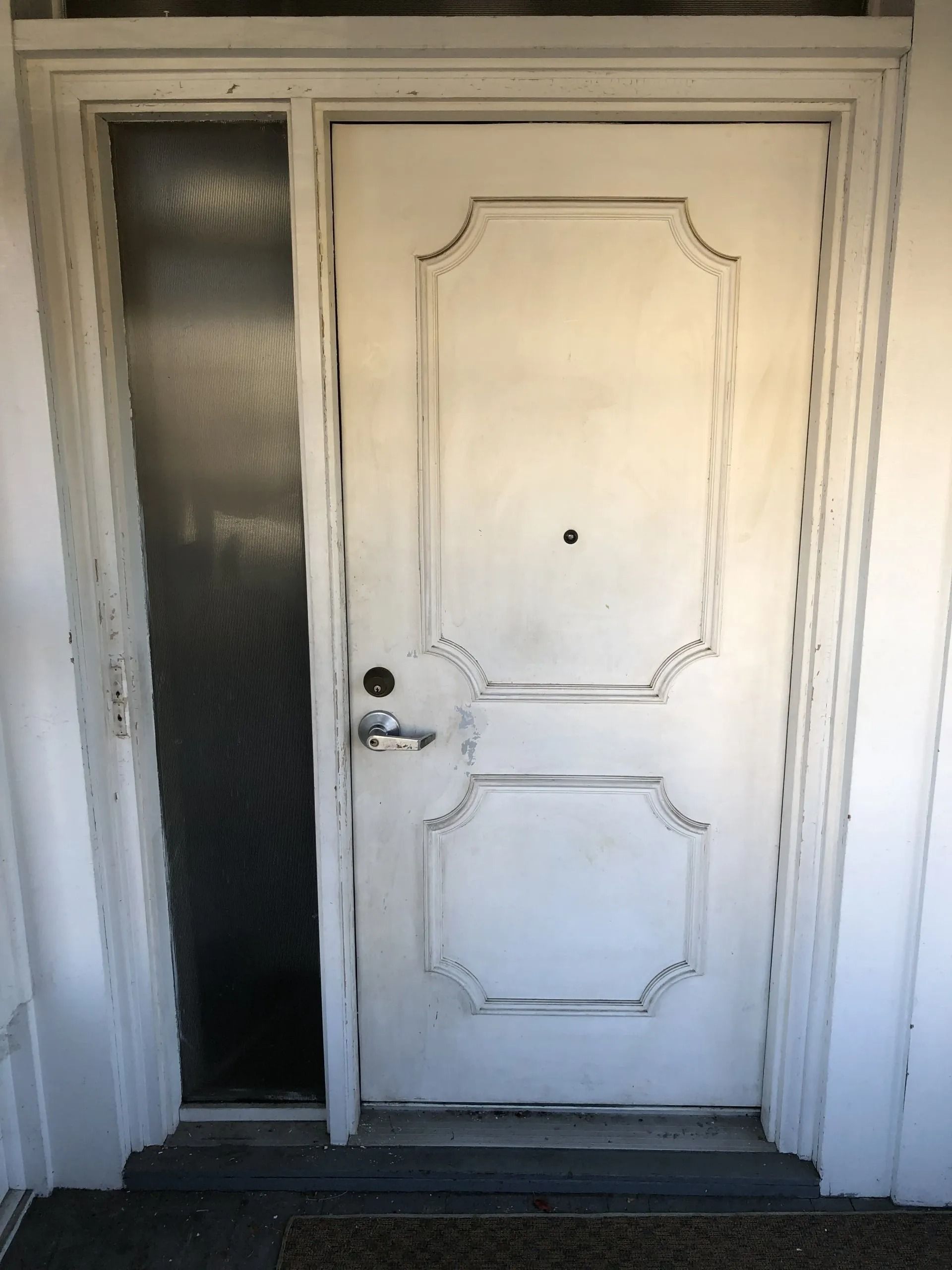 White front door with frosted glass side panel and decorative molding, slightly worn paint.