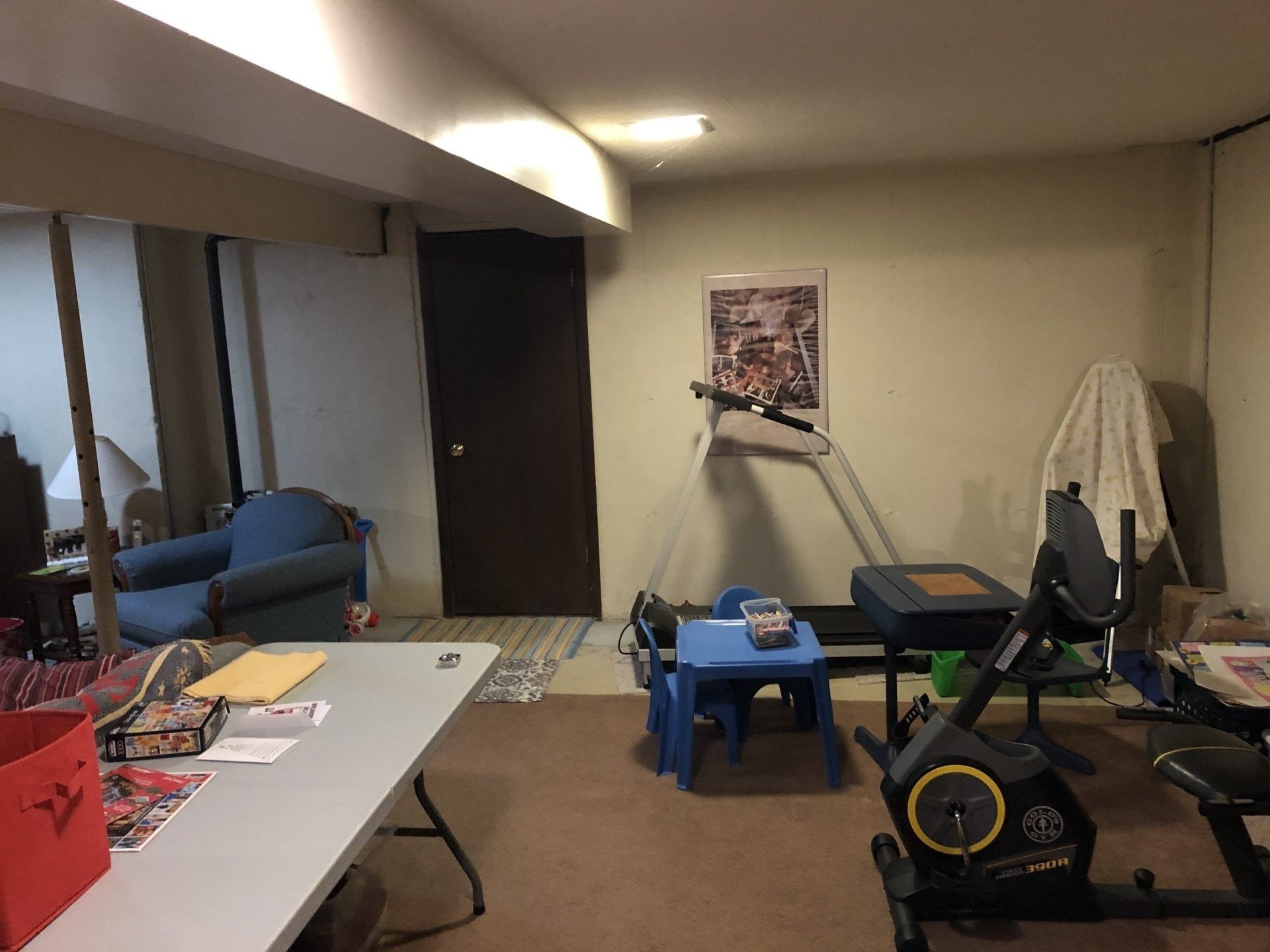 Basement room with a table, treadmill, exercise bike, blue stool, and a windowed wall.