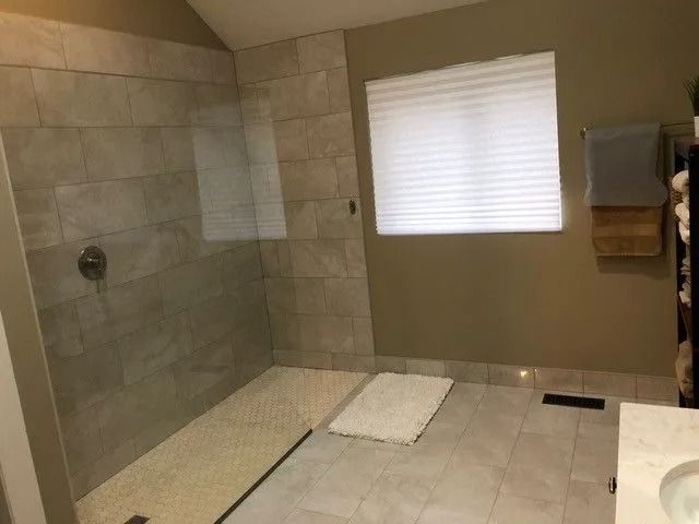 Modern bathroom with tiled walk-in shower, glass partition, window blind, and two floor mats