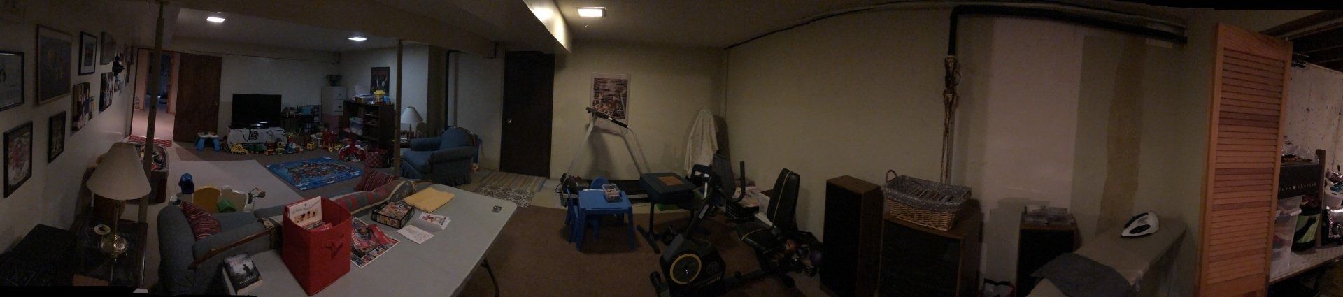 Dim indoor room with tables, chairs, and scattered items in a cluttered workspace or storage area.