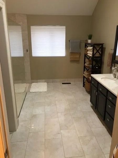 Empty beige bathroom with tiled floor, vanity sink, window blinds, shower at left, and shelving unit at right