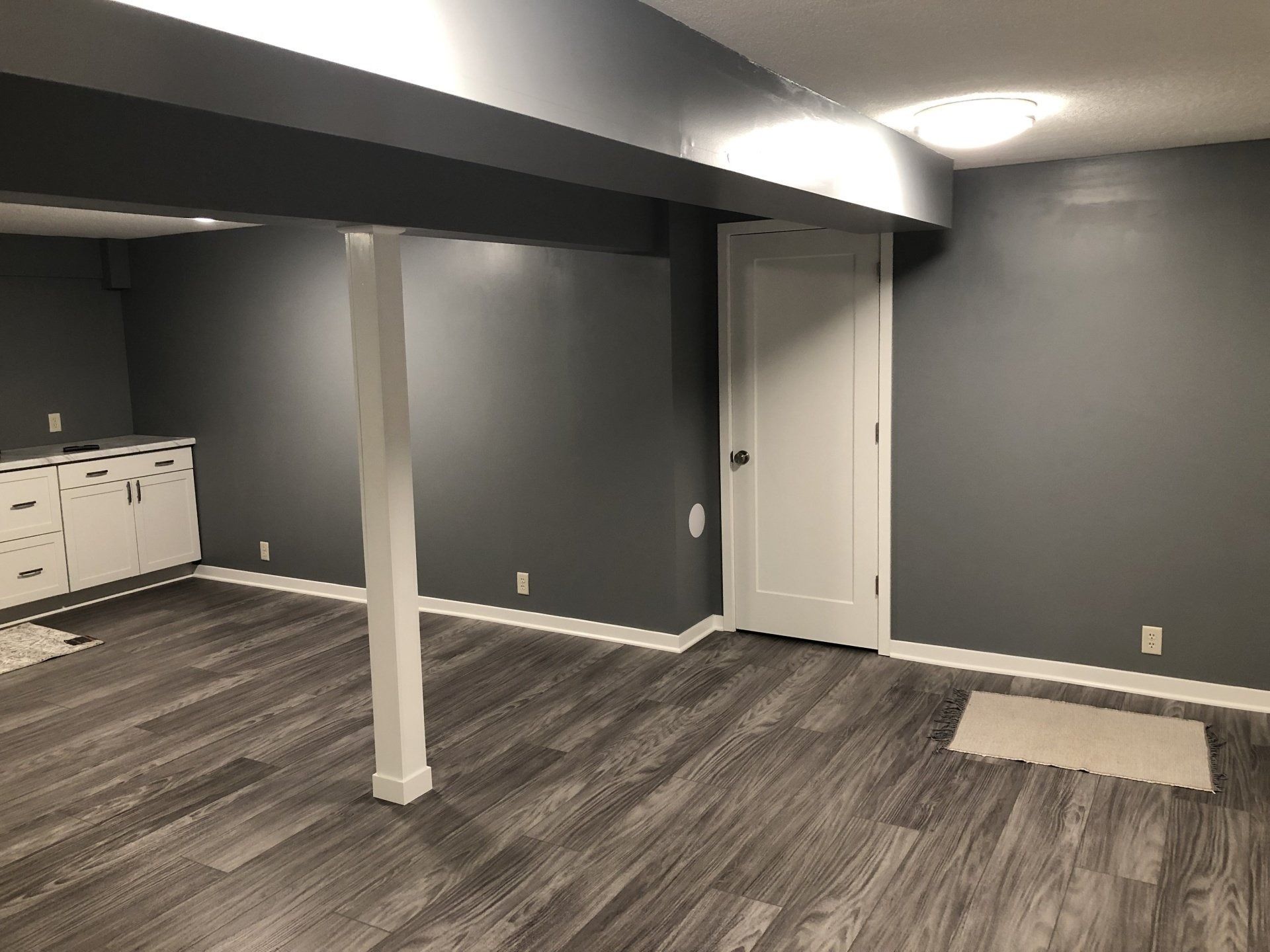 Empty gray basement room with white columns, wood-pattern floor, and a white door.