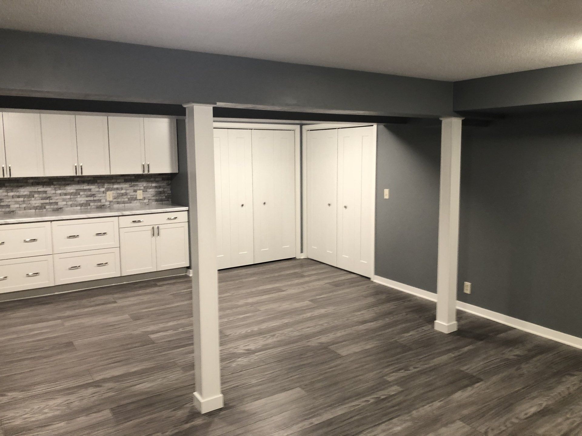 Empty gray basement room with white cabinets, closet doors, and two support columns