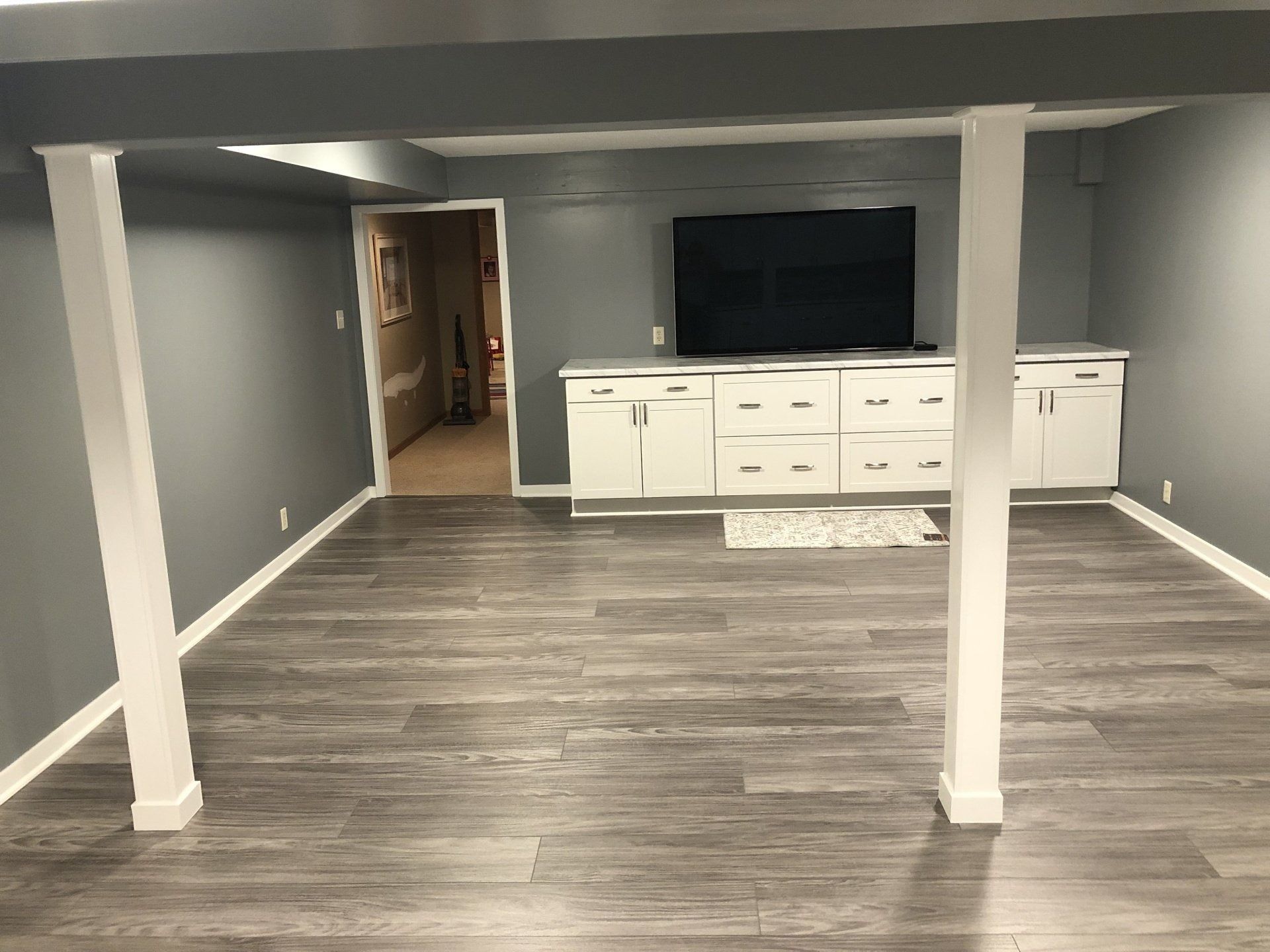 Basement family room with gray walls, wood flooring, white support posts, and a TV on a white cabinet