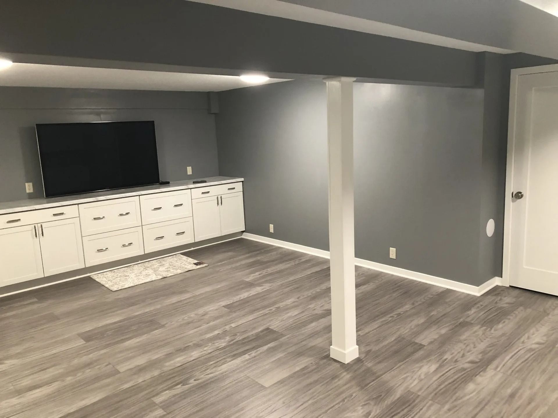 Finished basement room with gray walls, wood flooring, white cabinets, a TV, and a support column.