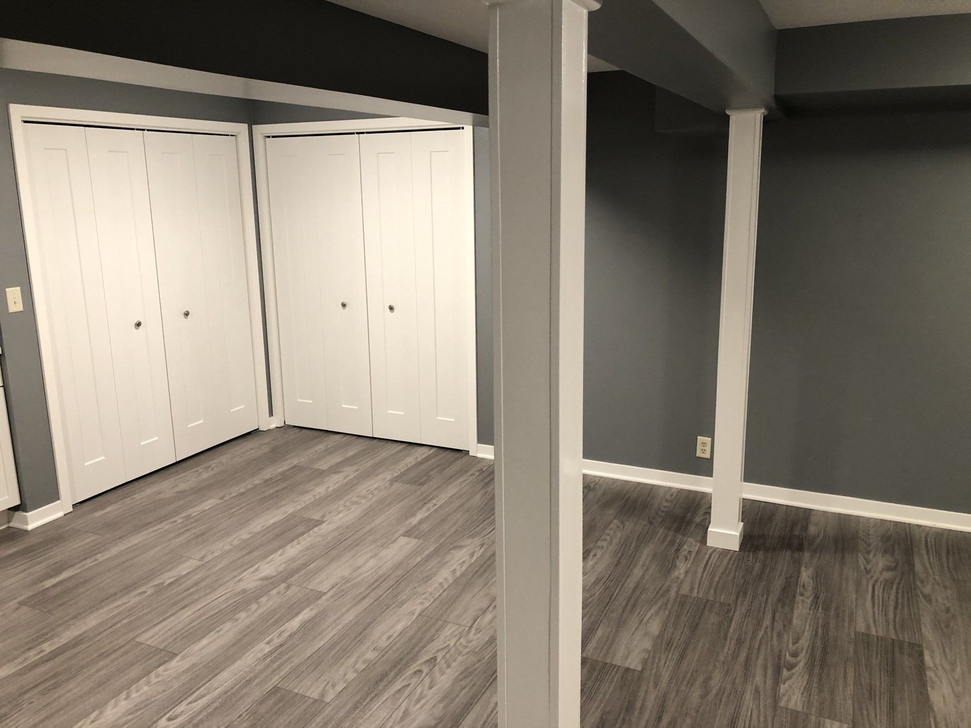 Empty basement room with gray walls, white support posts, and white closet doors on wood-look flooring