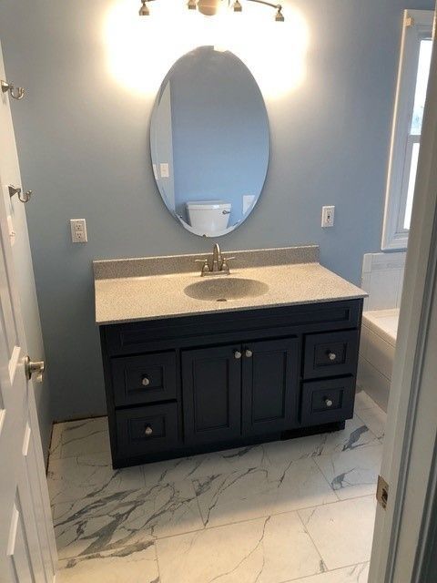Modern bathroom vanity with black cabinets, white countertop, oval mirror, and marble-look tile floor.