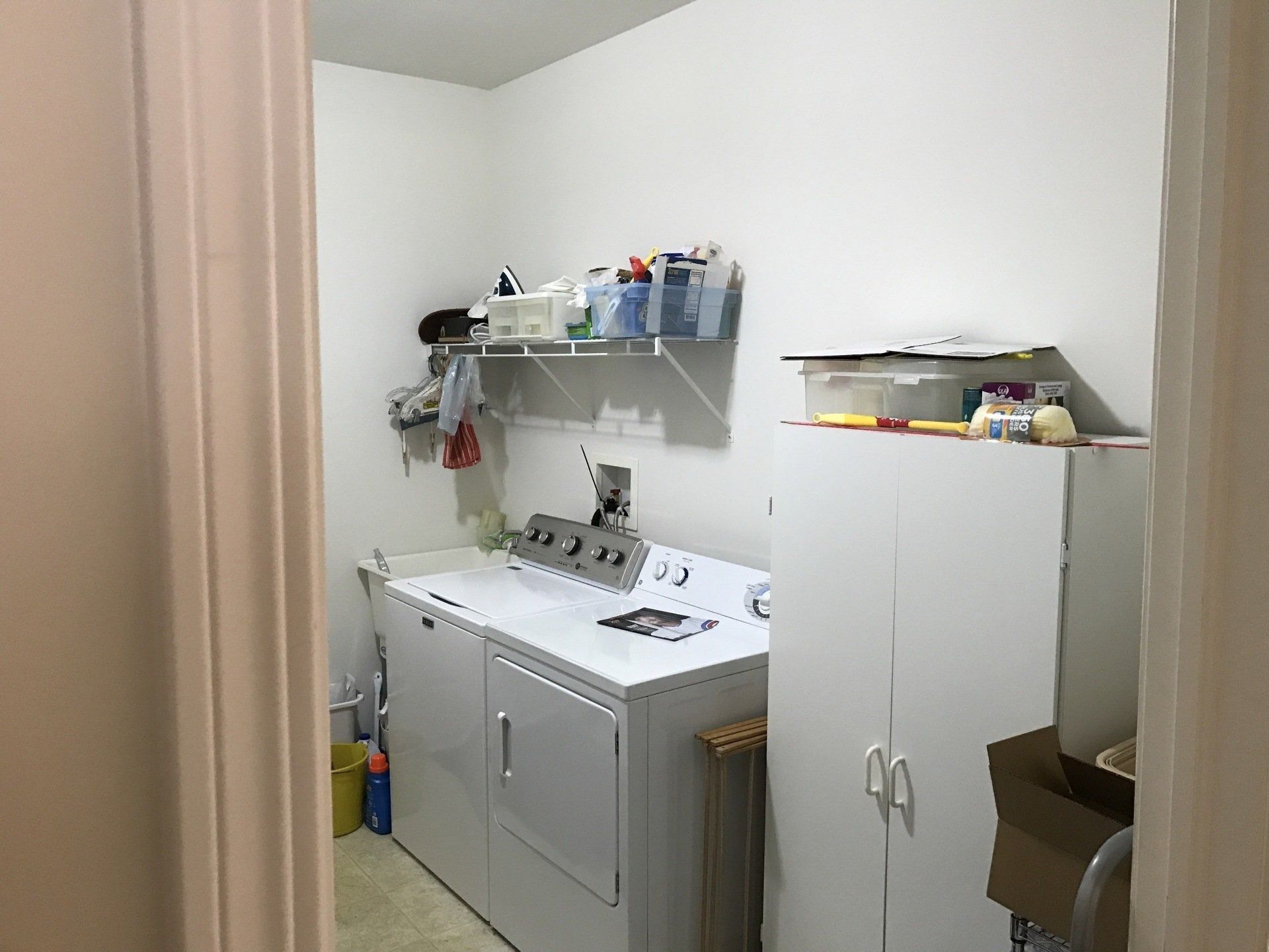Laundry room with a white washer and dryer, wall shelf, and cabinets beside a doorway.
