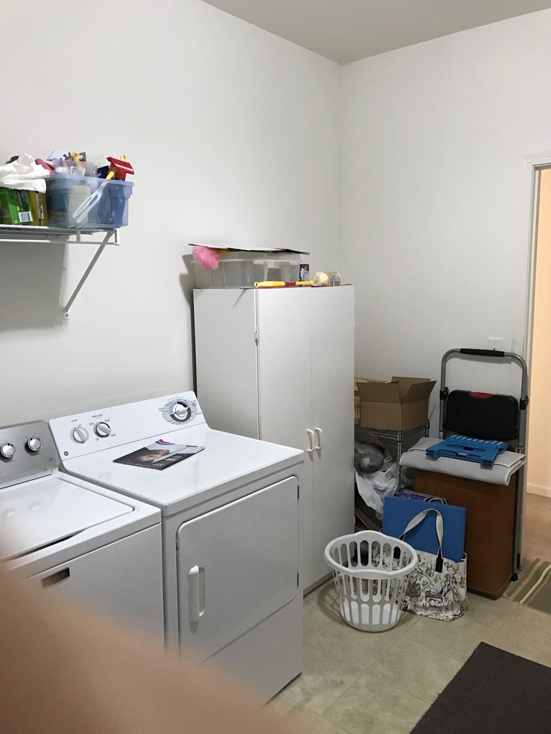 Laundry room with white washer and dryer, storage shelves, and a white cabinet against plain walls.