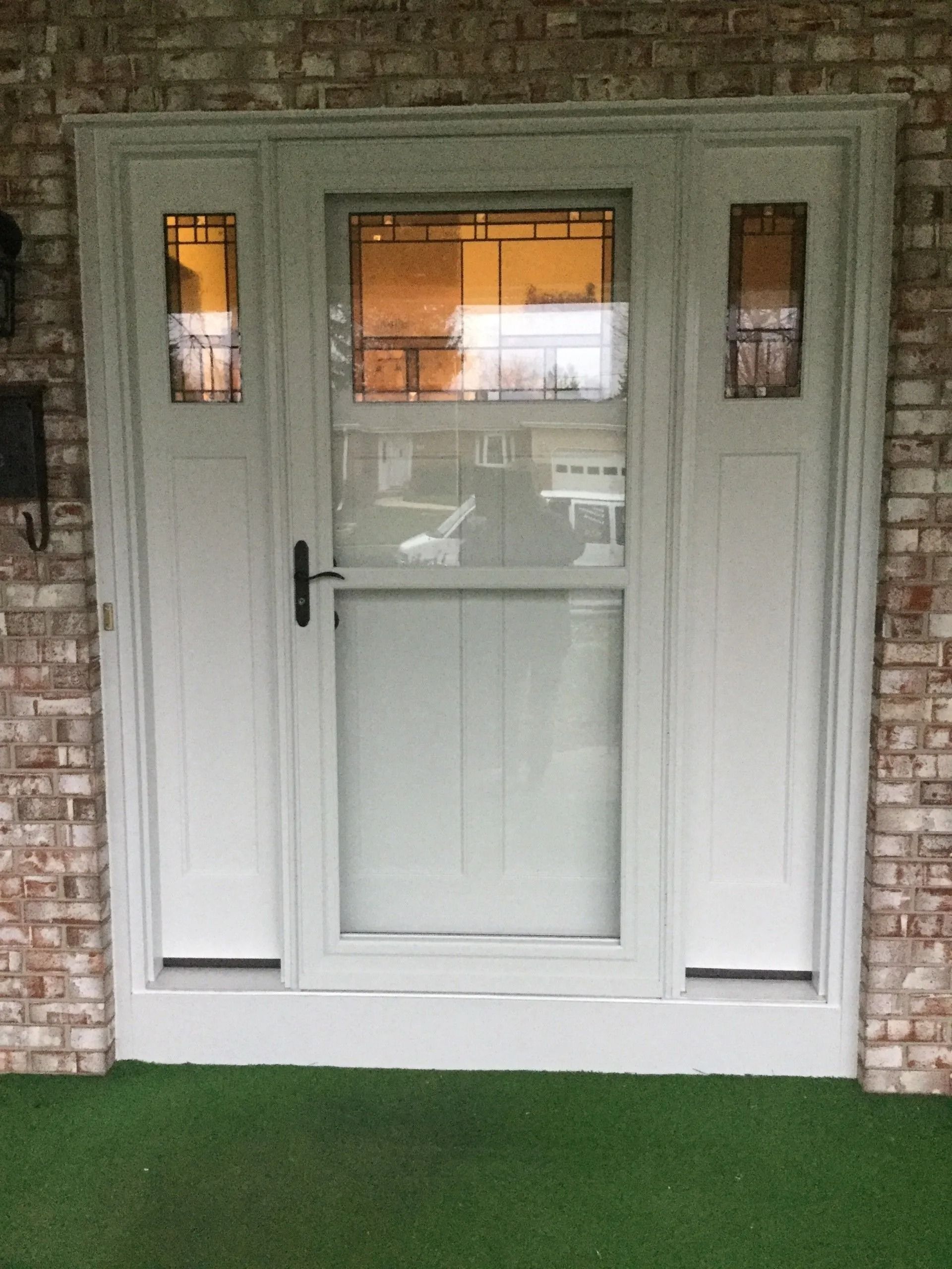 White front door with glass panes and sidelights set in a brick wall, above green artificial turf