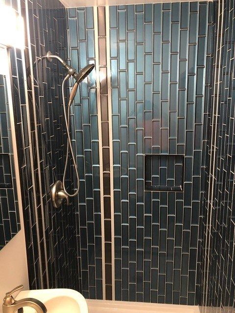 Modern shower with dark blue tile, handheld showerhead, glass door, and sink edge visible