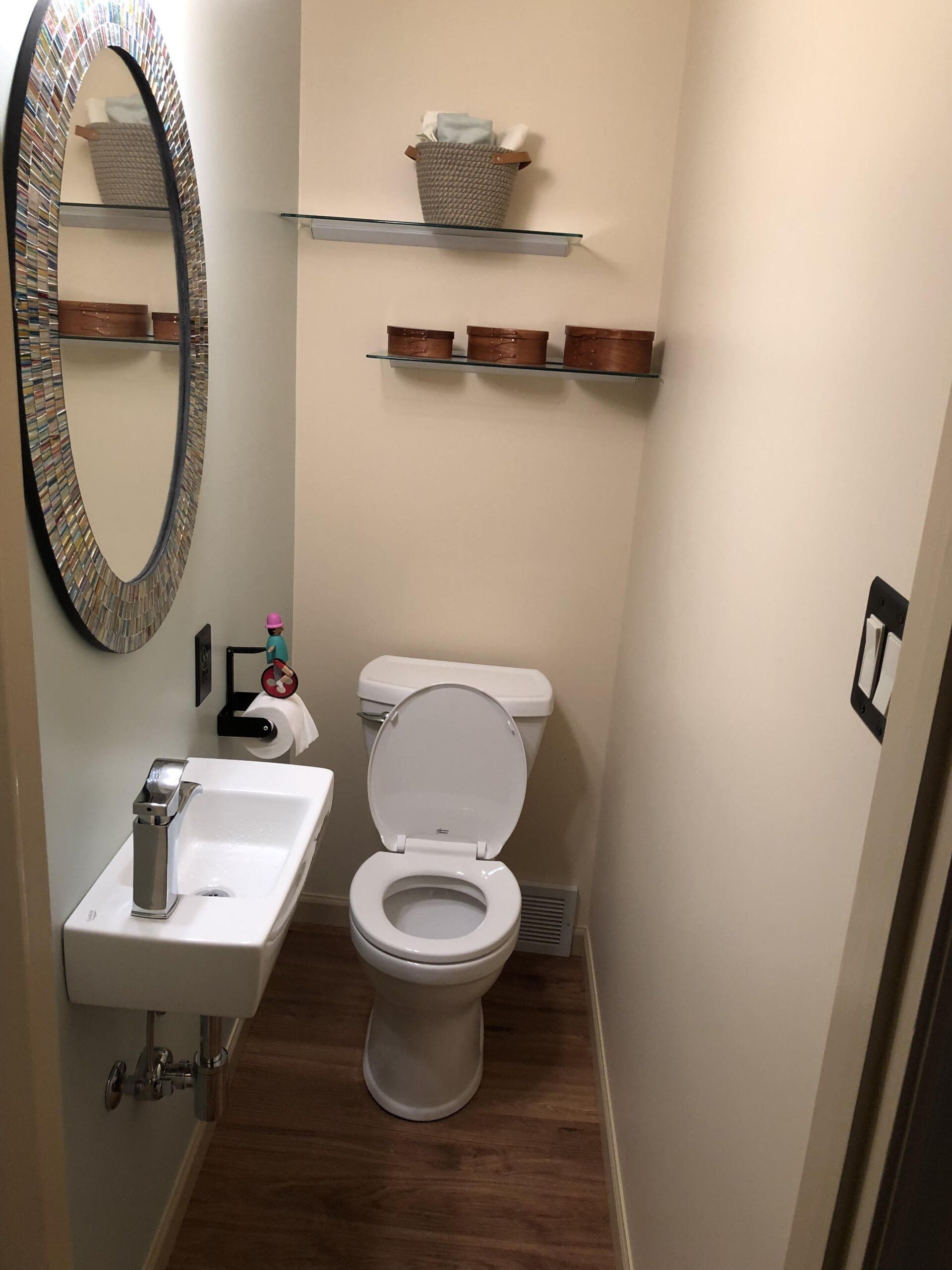 Small beige bathroom with sink, toilet, oval mirror, wall shelves, and wood-look floor.