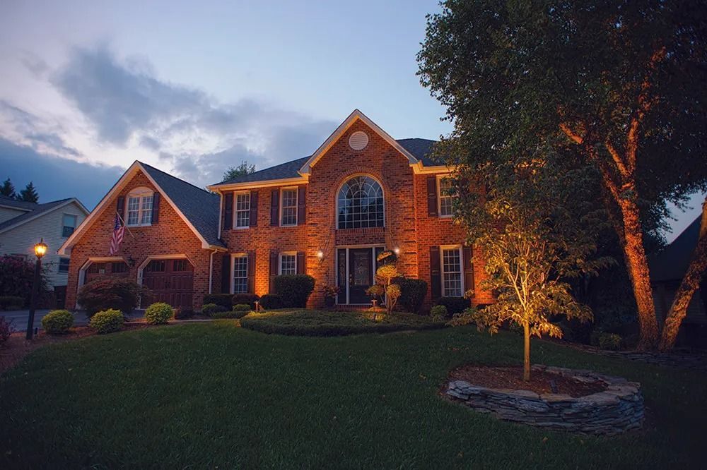 Brick house at dusk with lit windows, a large arched entry, and a tree-lit front lawn.