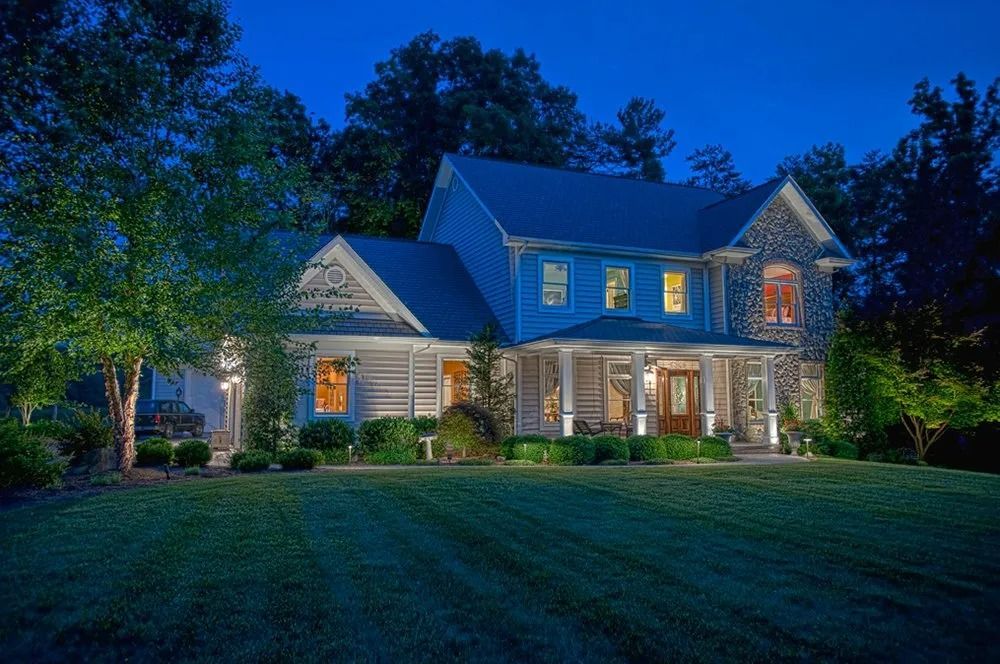 Large two-story house with lit windows and porch at dusk, surrounded by trees and a green lawn.