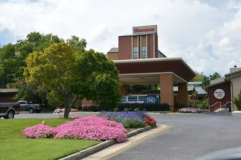 Hotel entrance with brick portico, landscaped flowers, and driveway under a cloudy sky