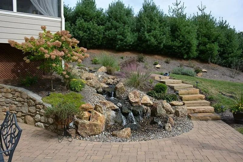 Landscaped backyard with stone waterfall, steps, shrubs, and brick patio beside a house
