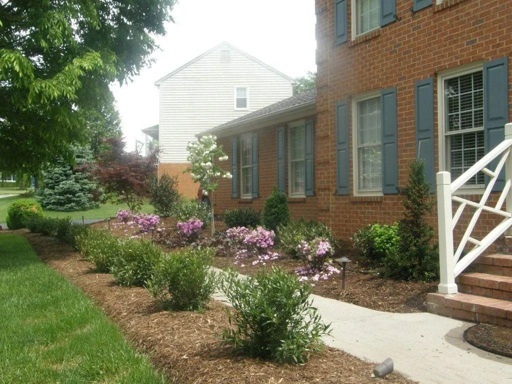 Brick house front walk with flower beds, green shutters, and a white porch railing