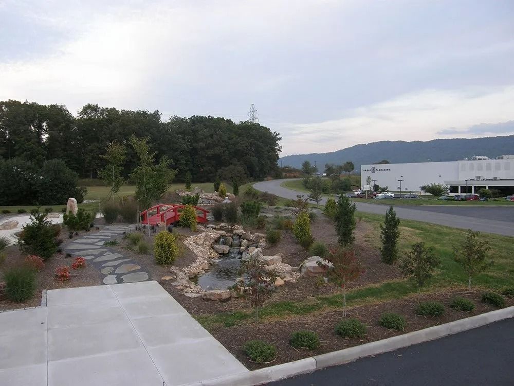 Landscaped garden with stone path, small pond, trees, and a white building near a road and hills.