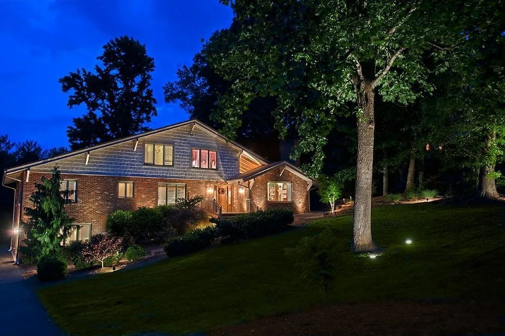 Well-lit house at night surrounded by trees and a sloping lawn under a deep blue sky