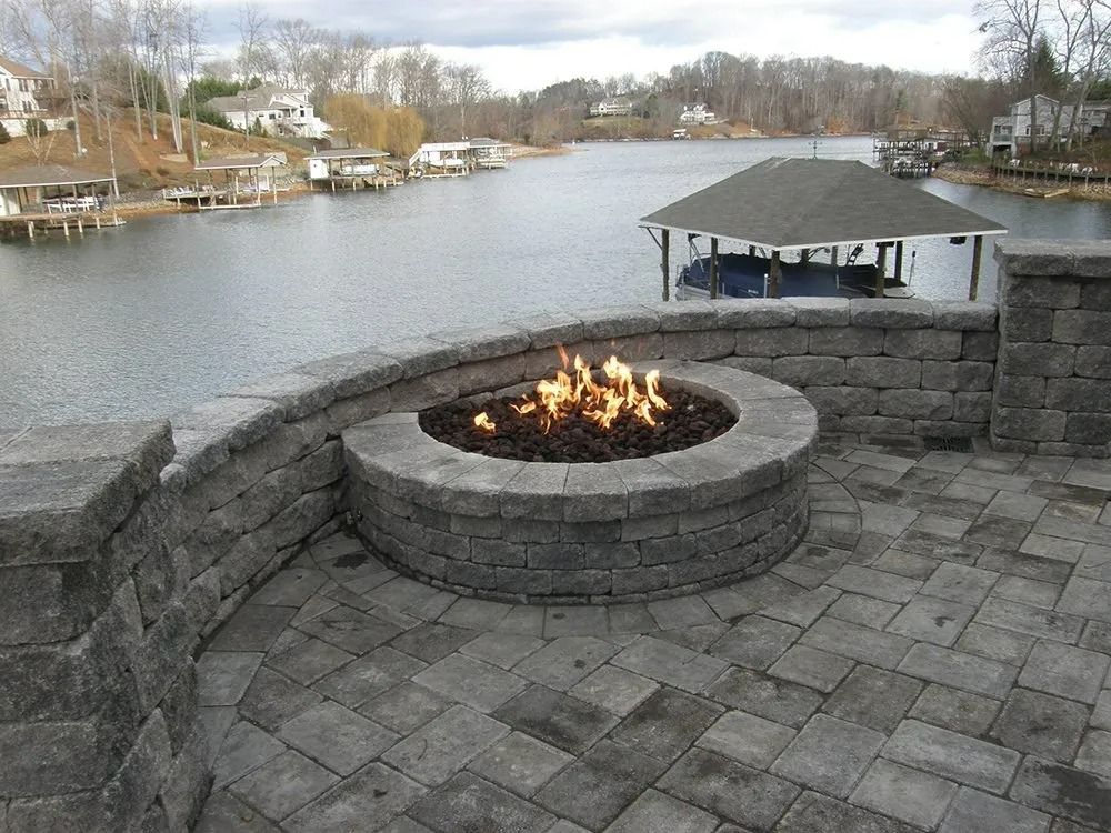Stone patio with circular fire pit overlooking a calm waterfront and distant houses