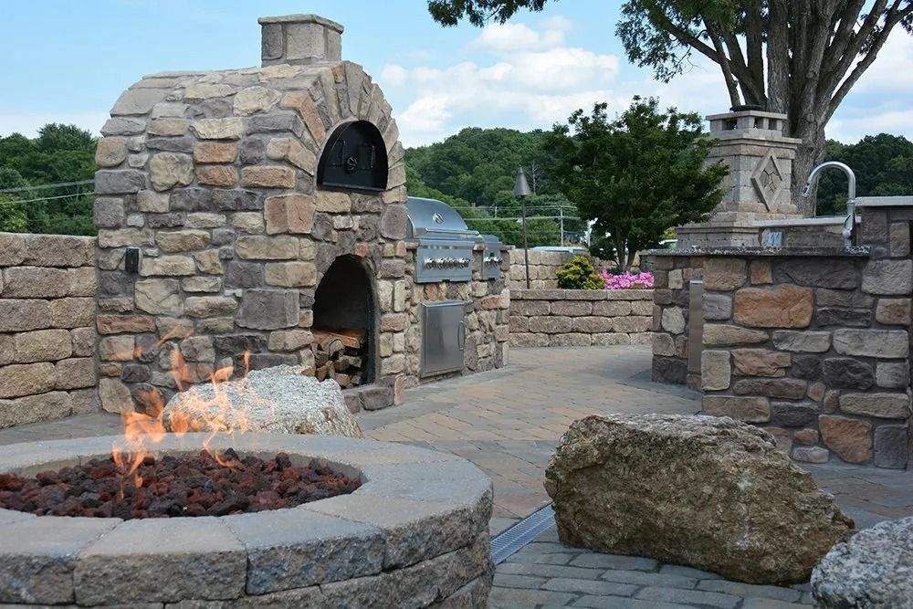 Stone outdoor fireplace with burning fire beside a paved patio and landscaping