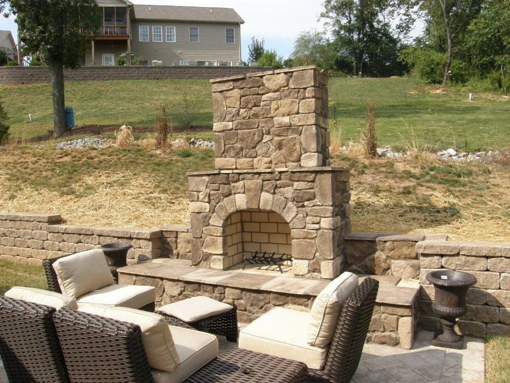 Stone outdoor fireplace with wicker patio seating in a backyard, with a grassy slope and house in the background