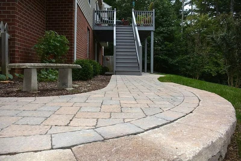 Curved stone walkway beside a brick building, with stairs and a bench in a landscaped courtyard