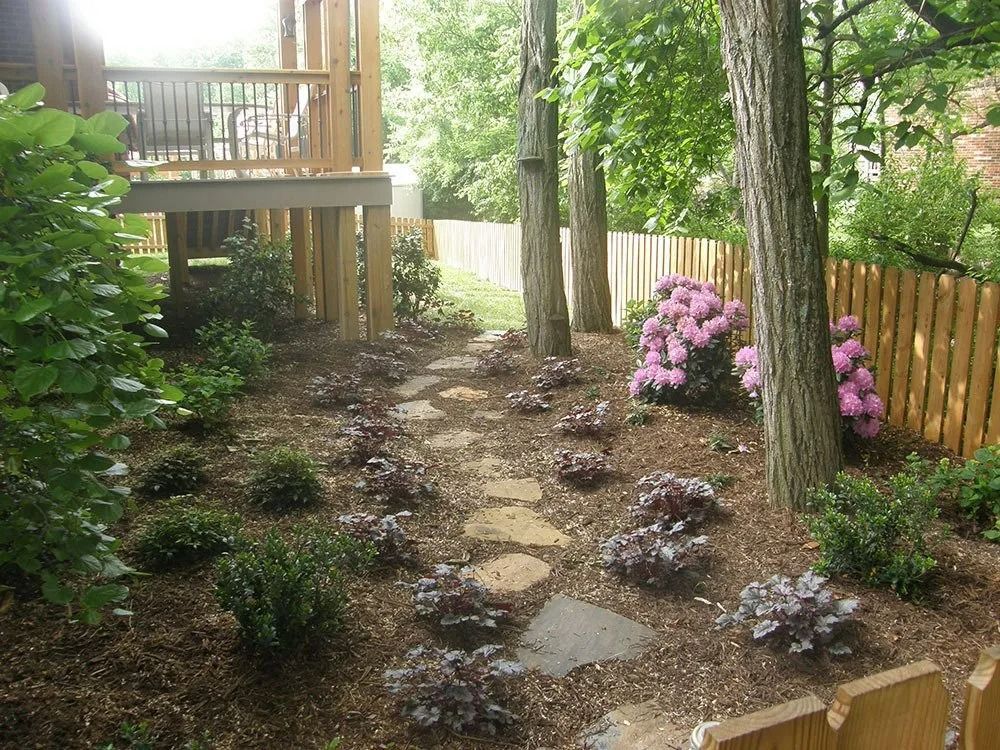 Garden path with stepping stones, trees, and pink flowers beside a wooden fence and deck