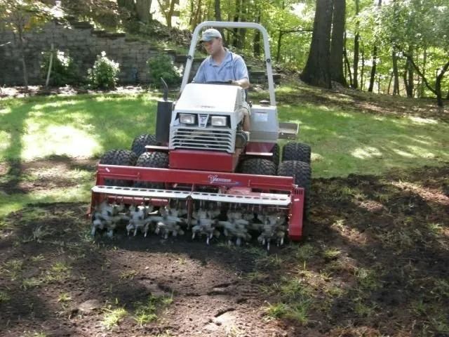 Red lawn tractor mulching leaves on a grassy, wooded slope with an operator seated inside the cab