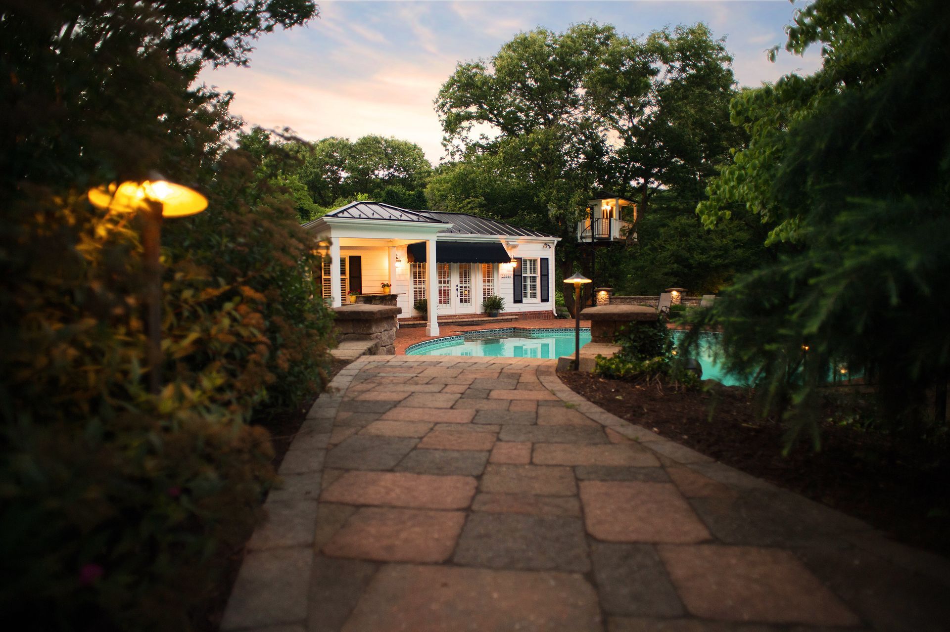 Stone path leading to a lit house and pool at dusk, surrounded by lush trees and garden lights