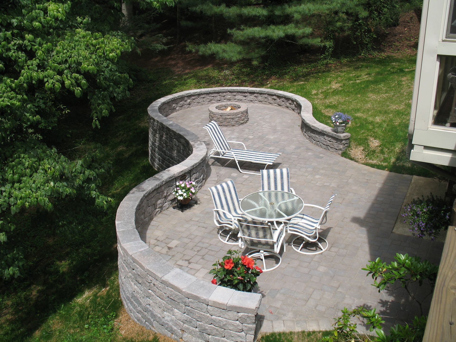 Stone patio with striped chairs, table, and flower pots in a landscaped backyard