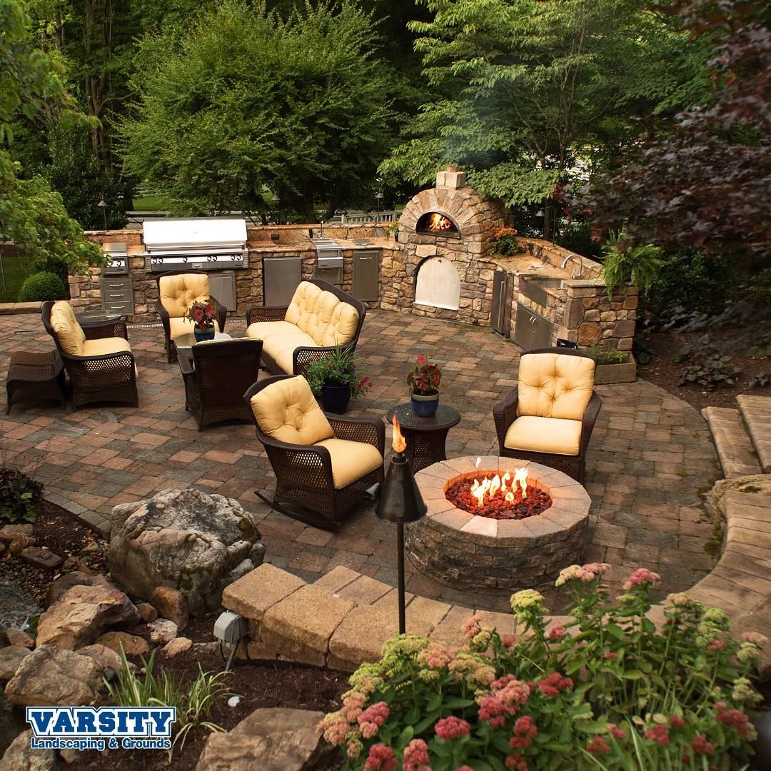 Backyard stone patio with cushioned chairs and a fire pit surrounded by trees and flowers
