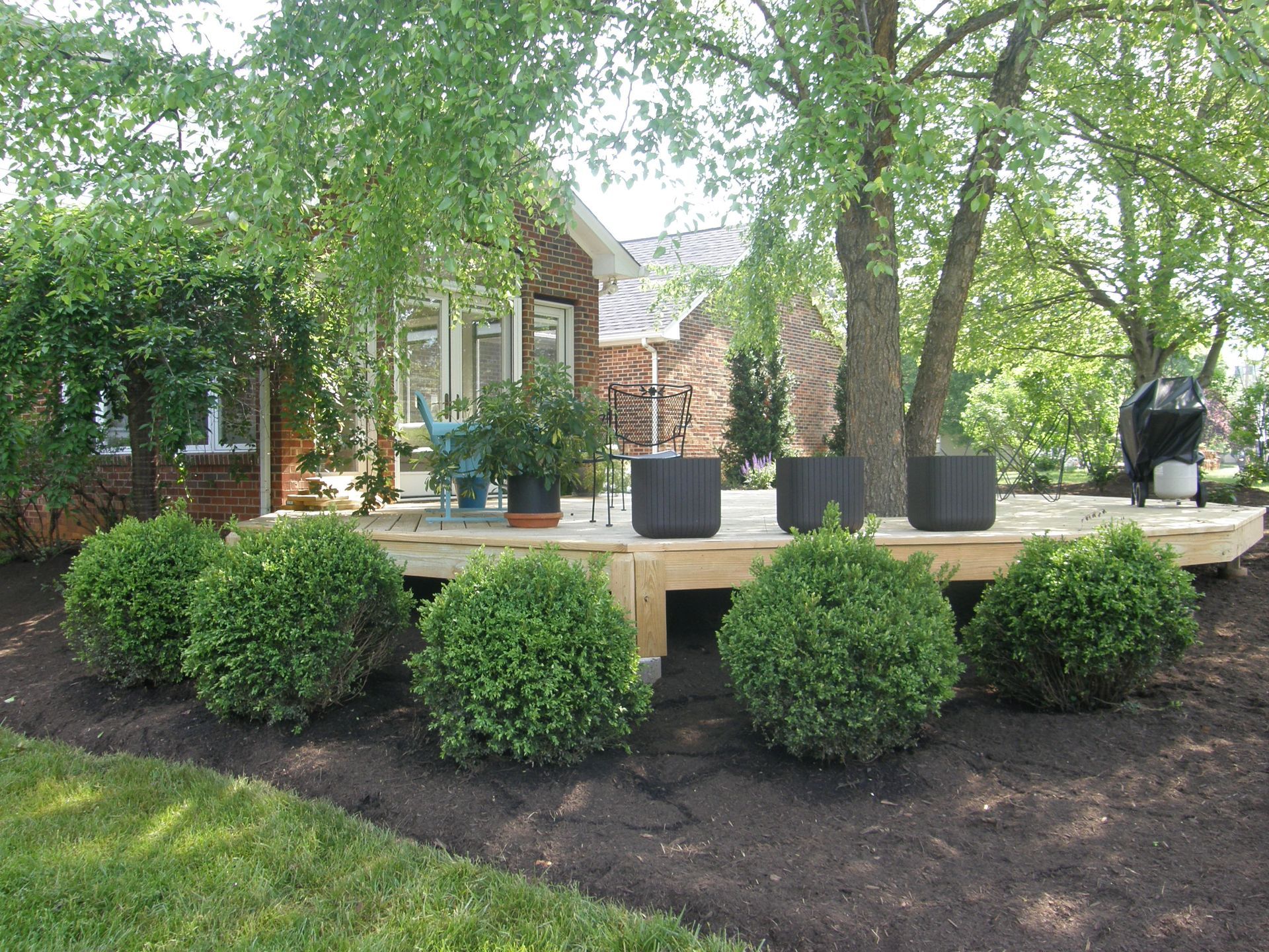 Front yard patio with potted plants, trimmed shrubs, and a brick house under leafy trees