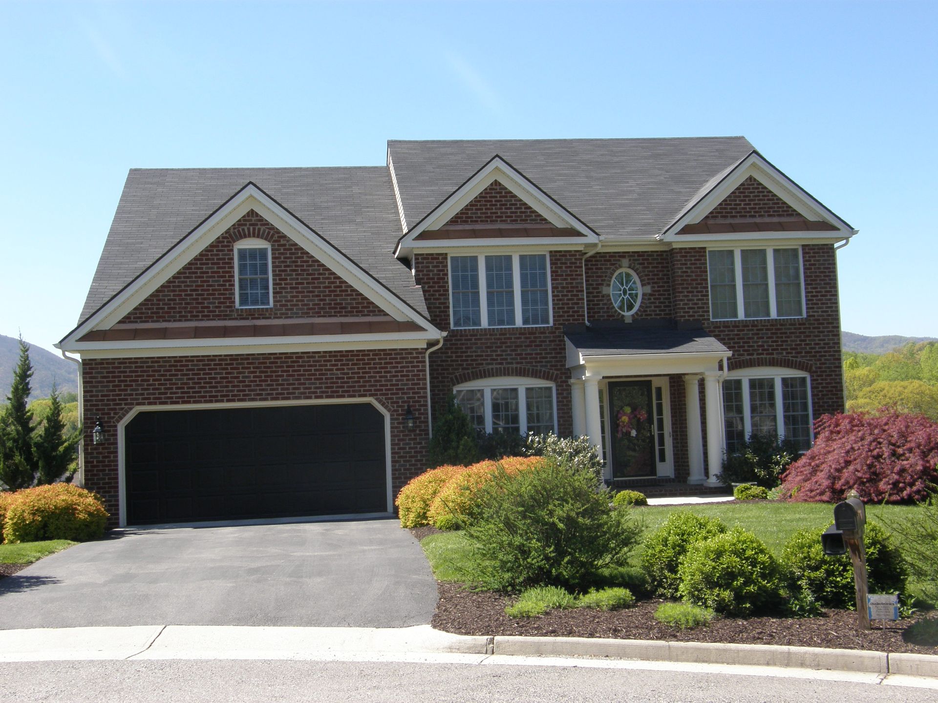 Two-story brick suburban house with dark roof, black garage door, and landscaped front yard